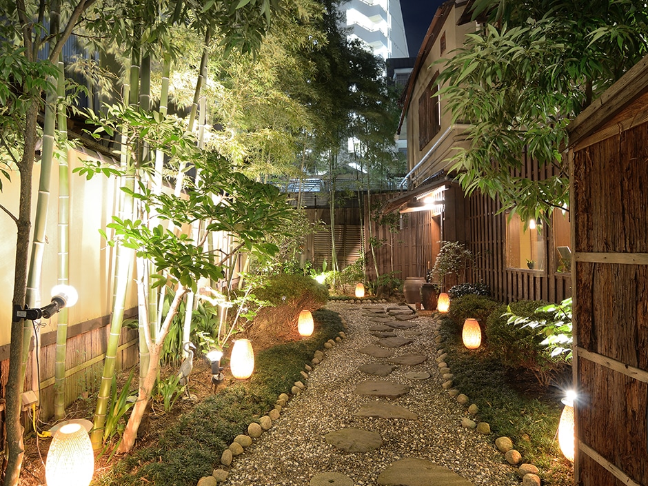 Bamboo trees and gravel path lined with lanterns in a Japanese garden at night.