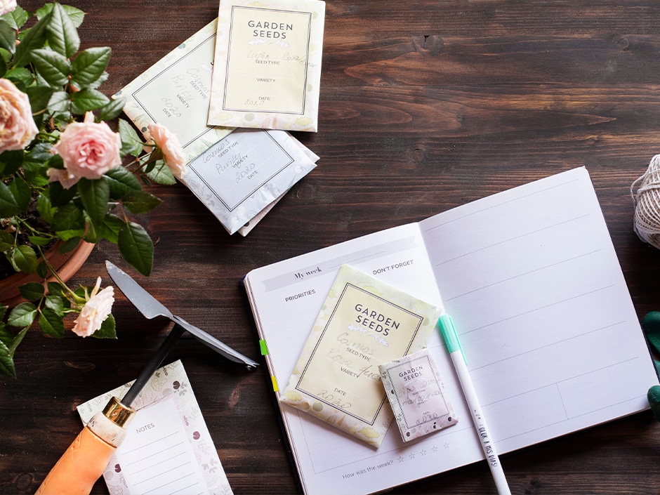 A gardening planning scene on a wooden table featuring an open planner, various garden seed packets, a pair of scissors, twine, and fresh flowers.